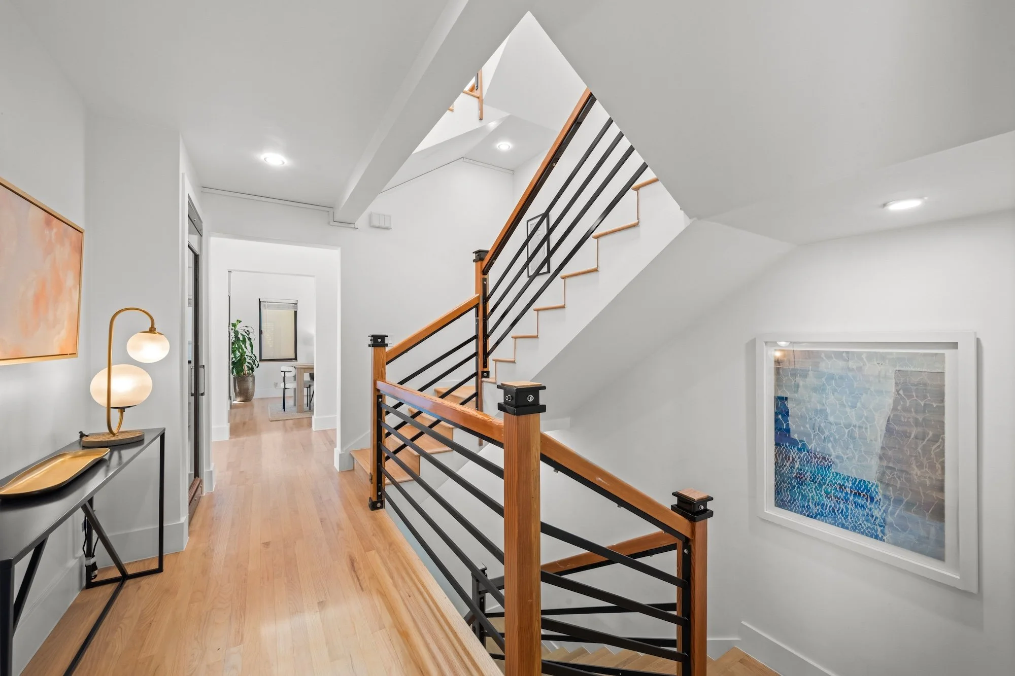 Interior view of a modern home with white walls and wooden flooring. A staircase with wooden handrails and black metal balusters leads upstairs. There is a decorative lamp, a framed artwork, and a console table with a tray on it on the left side. An 