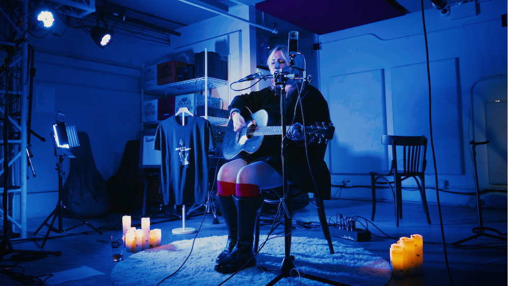 A woman with blonde hair plays guitar and sings into a microphone during a live performance in a dimly lit room with blue lighting, with candles and a T-shirt on a hanger as decoration.