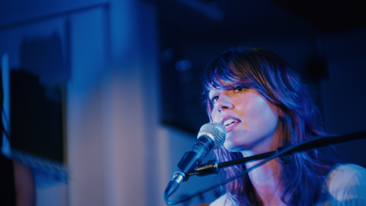 Female musician singing into a microphone while playing a keyboard in a dimly lit room with blue lighting.
