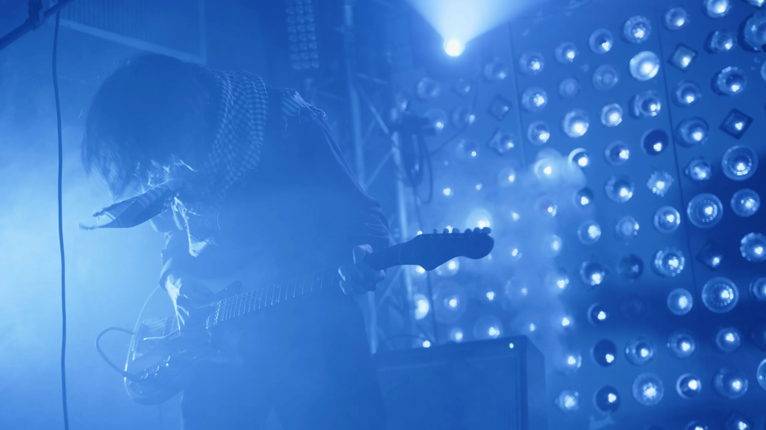 A musician playing an electric guitar on stage illuminated by blue and white stage lights in a dark setting.