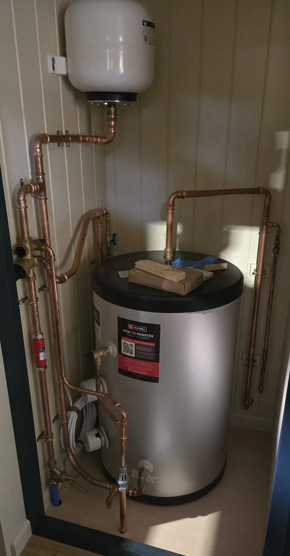 A water heater with exposed copper piping and a white expansion tank mounted above it in a small utility closet.