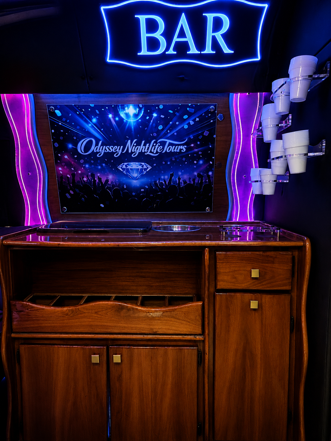 A bar area with a neon blue sign that says 'BAR' at the top. Below is a sign for Odyssey NightLife Tours featuring a disco ball, a crowd of dancing silhouettes, and a large diamond graphic, all illuminated with blue and purple lights. The bar counter is made of wood with a small sink and some empty cup holders on the right side.