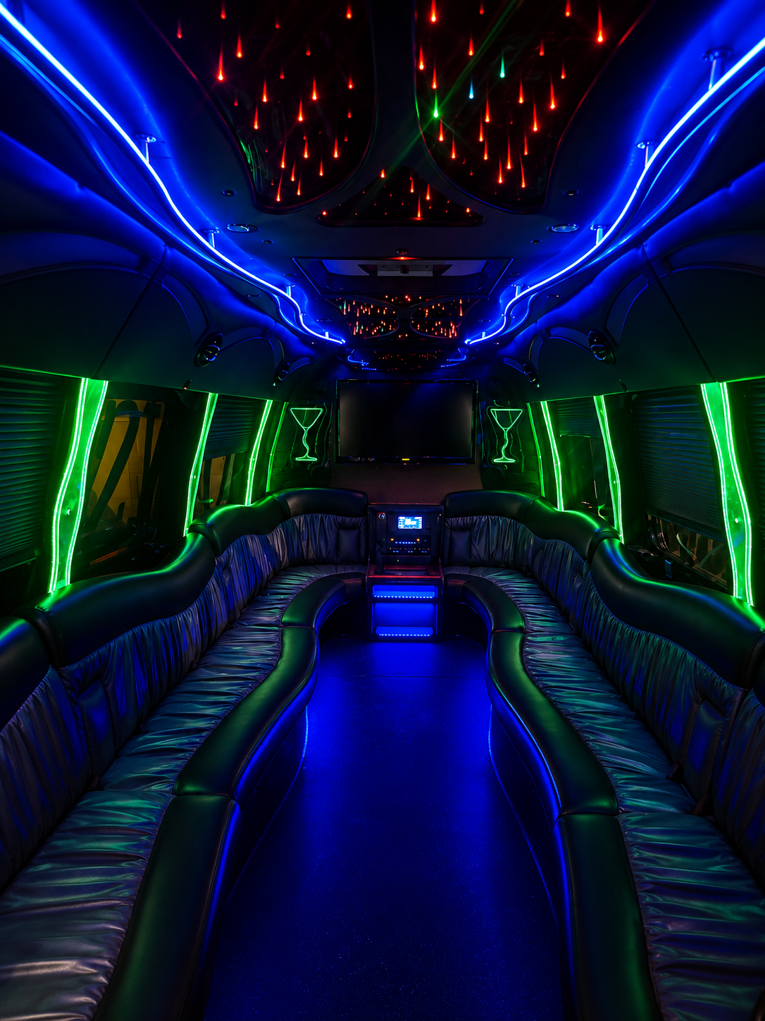 Interior of a party bus with black leather seating and neon lighting, including green and blue lights on the sides and neon red and green lights on the ceiling, with a large TV screen at the front.