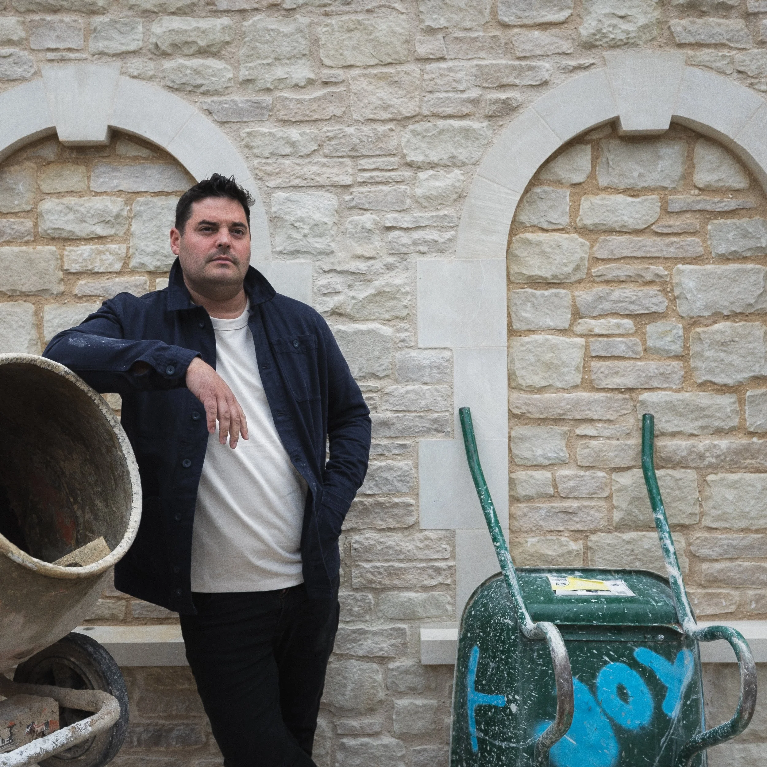 A man with short dark hair and a serious expression is standing outdoors beside a stone wall with arched windows. He is wearing a dark jacket over a white shirt. There are two green wheelbarrows nearby, with one of them partially visible.