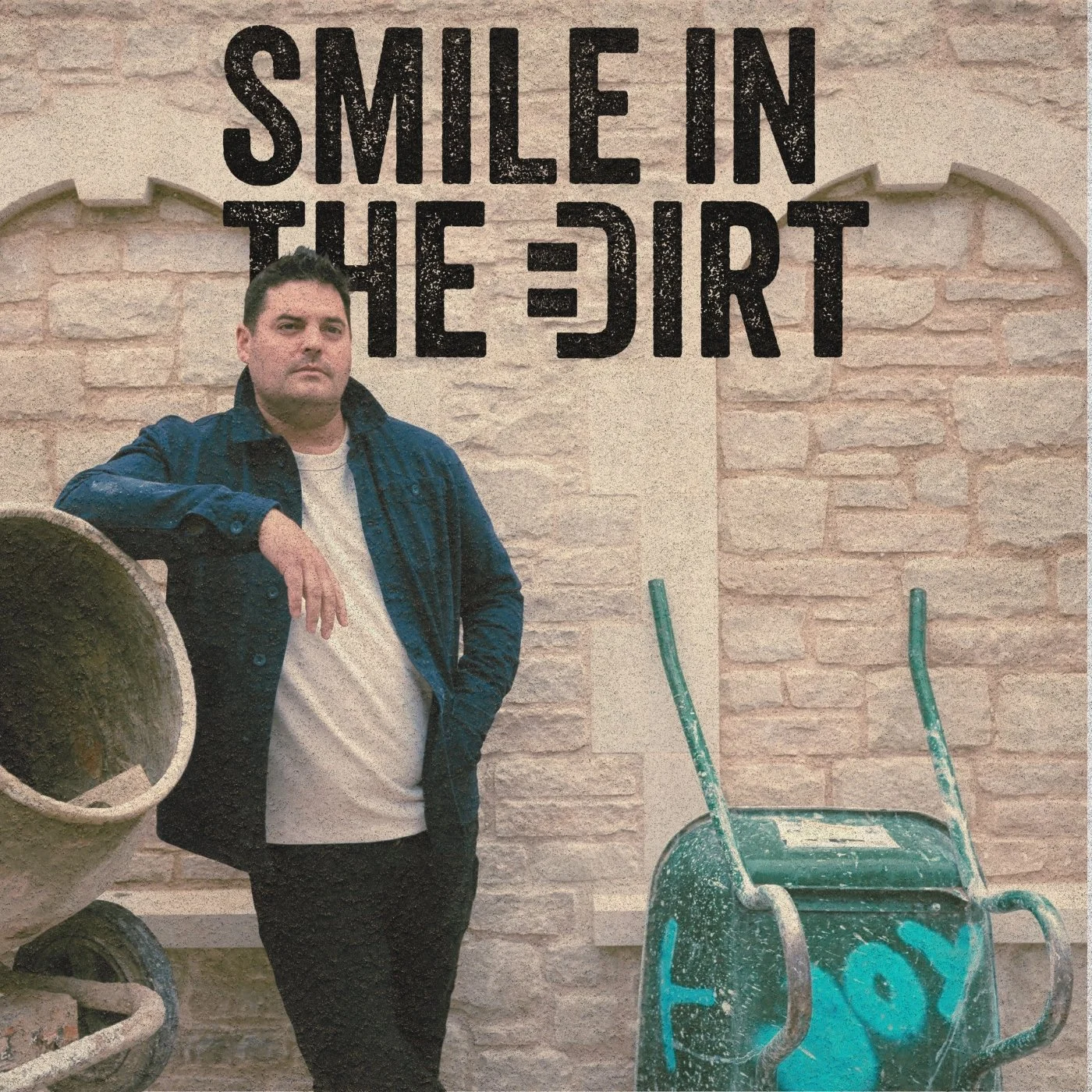 A man standing against a brick wall with the phrase 'Smile in the dirt' painted above. He is wearing a dark jacket over a light-colored shirt and has short dark hair. There are two green wheelbarrows in front of him, one with graffiti on it.