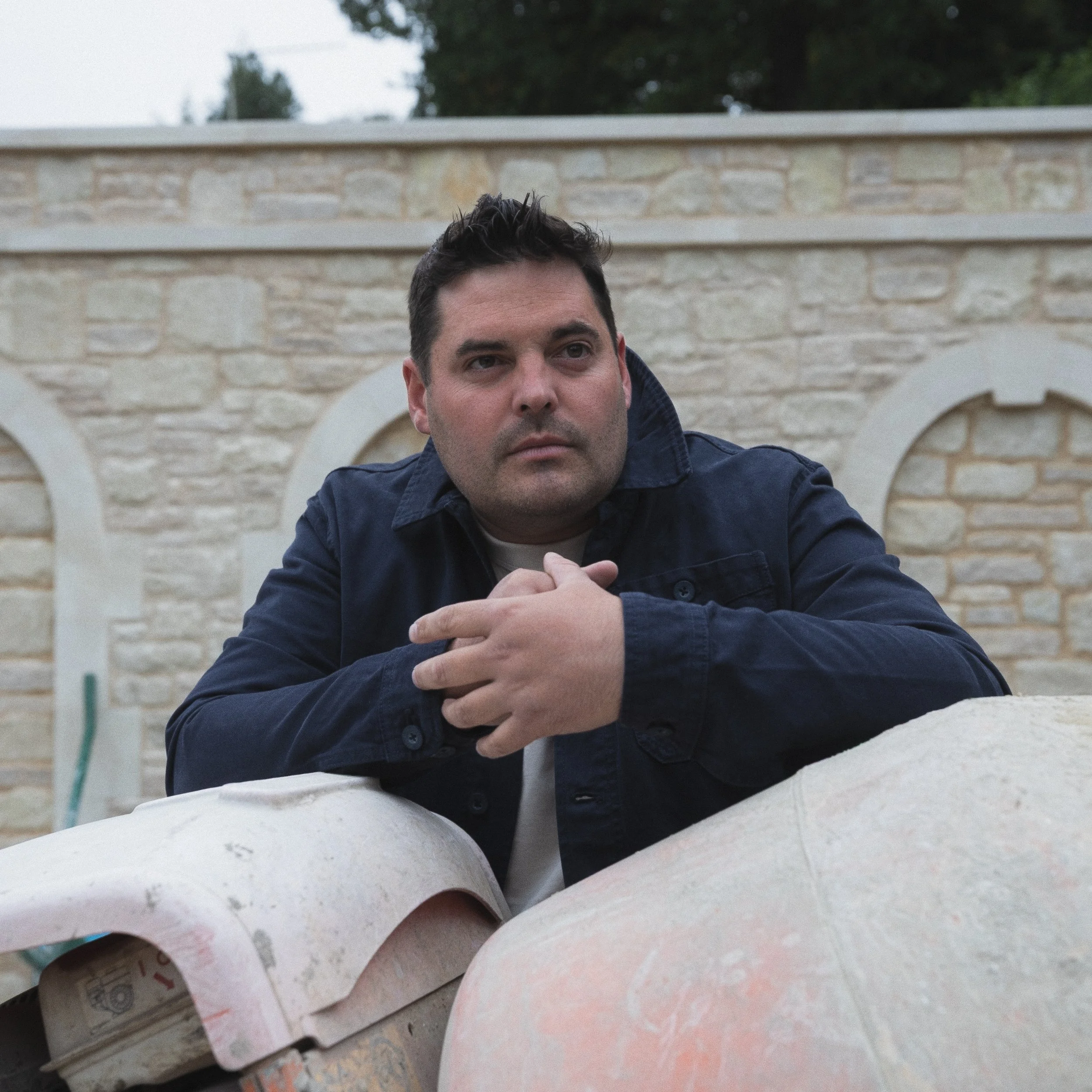 Close-up of a man with dark hair and a serious expression, resting his hands on a white stone surface, with a stone wall and trees in the background.
