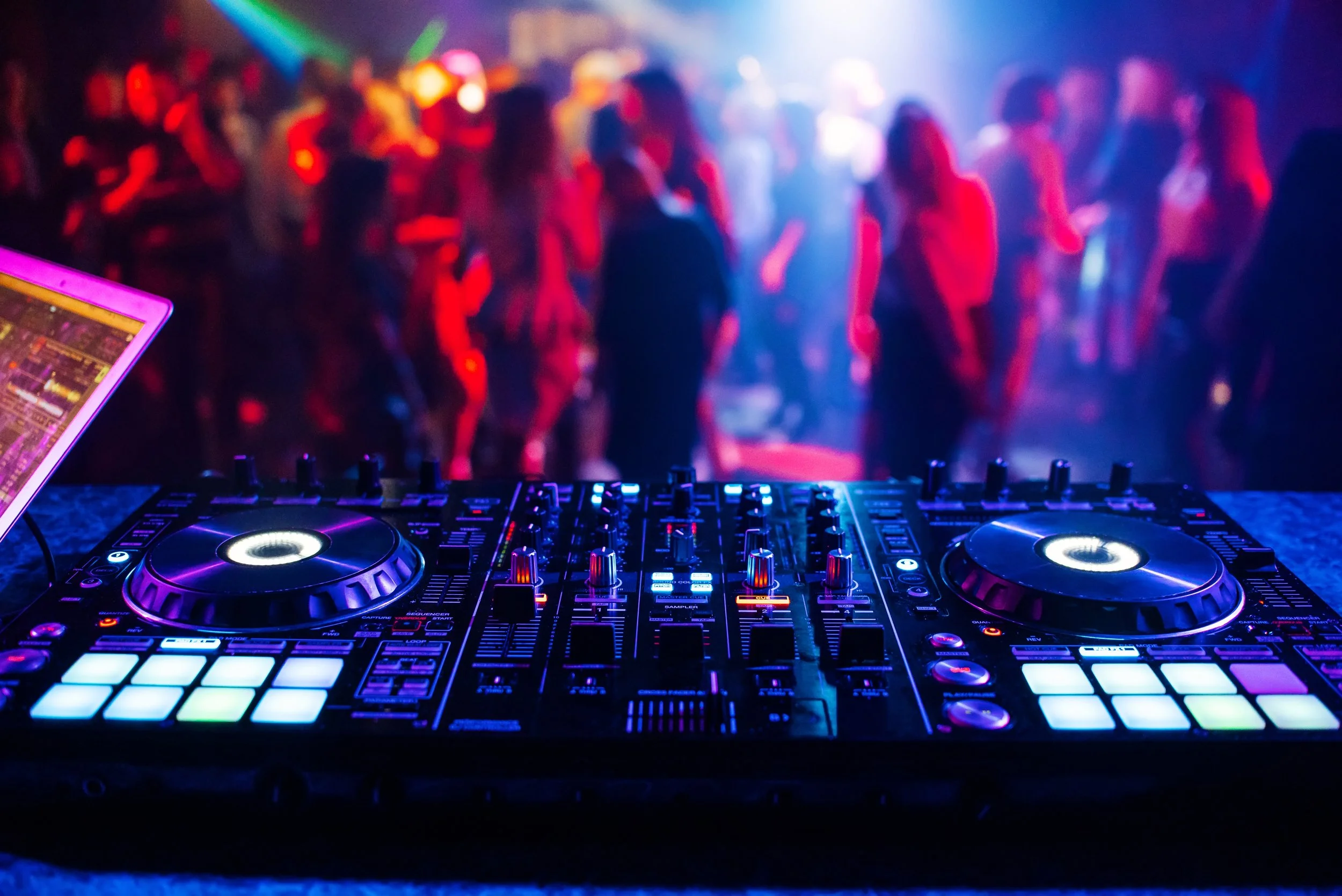 A DJ's mixing console with colorful LED lights in the foreground, and a blurred dance floor with people dancing under vibrant, multicolored lighting in the background.