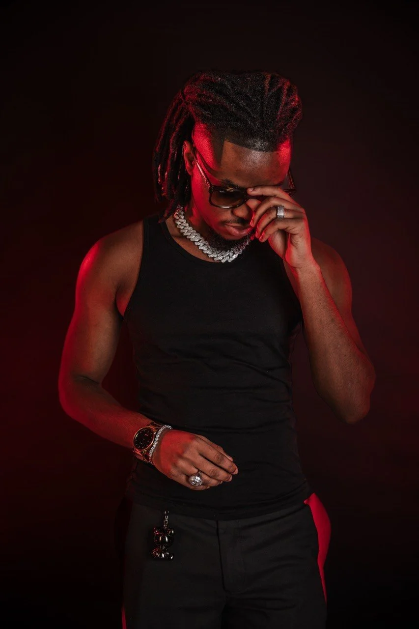 Man with dreadlocks wearing sunglasses, a silver chain necklace, a black tank top, and black pants with red accents, posing against a dark background with red lighting.
