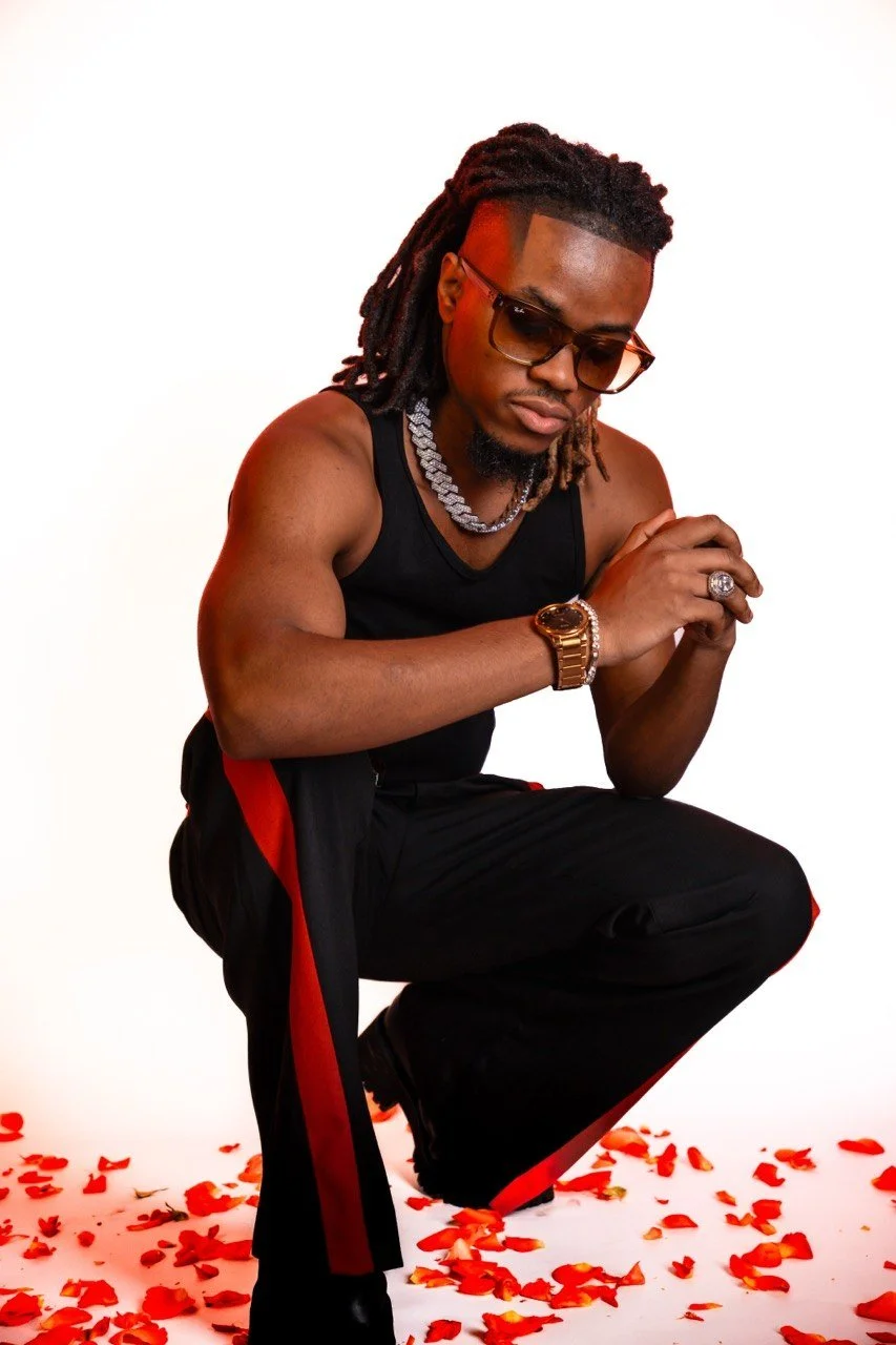A man with dreadlocks wearing sunglasses, a black tank top, black pants with red stripes, and gold jewelry, squatting among red rose petals on a white background.