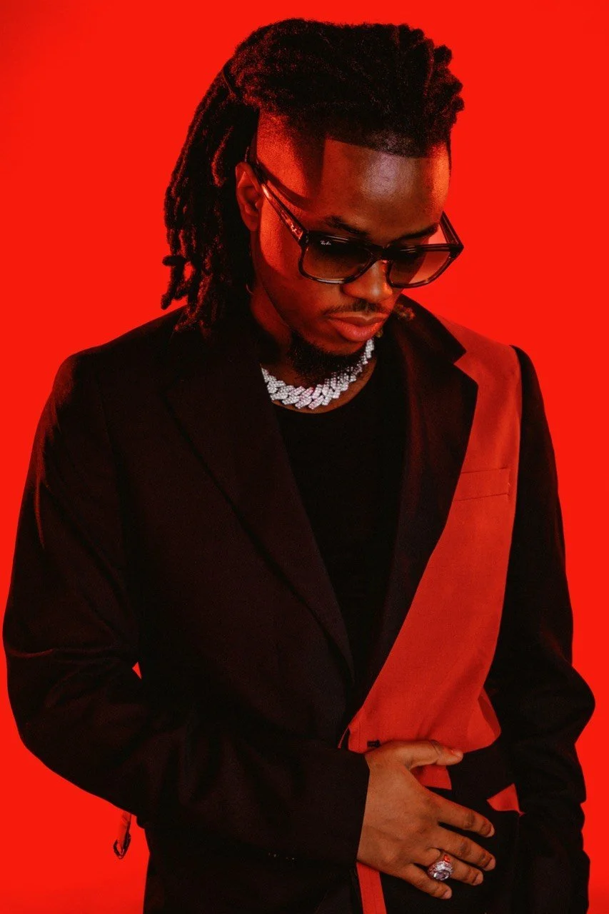 A man with dreadlocks wearing sunglasses, a black jacket with an orange accent, and a diamond necklace, standing against a red background