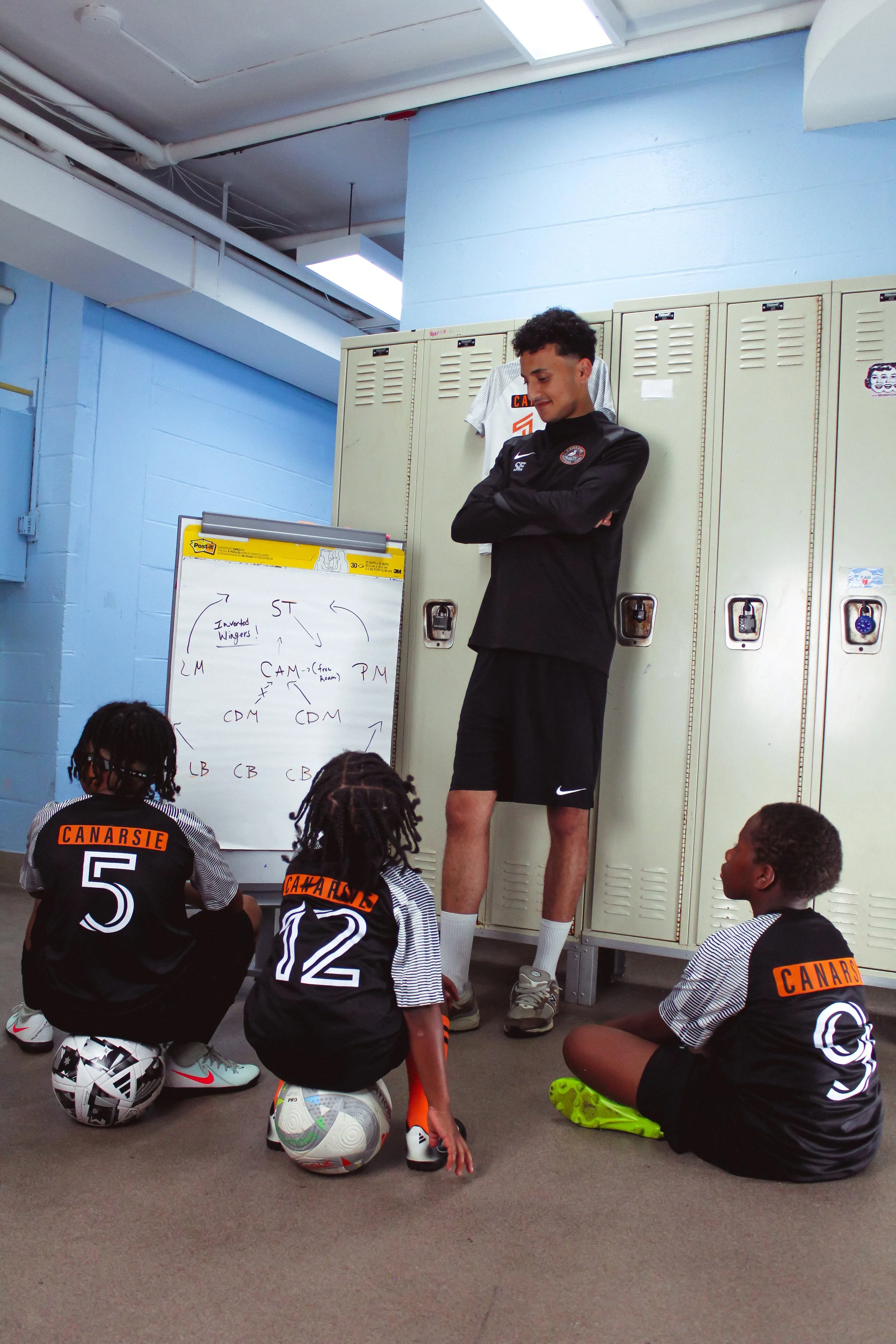 A coach standing in front of a row of lockers leading a classroom of young soccer players. The children are seated on the floor with soccer balls in front of them, listening intently. The scene takes place in a locker room or school hallway.