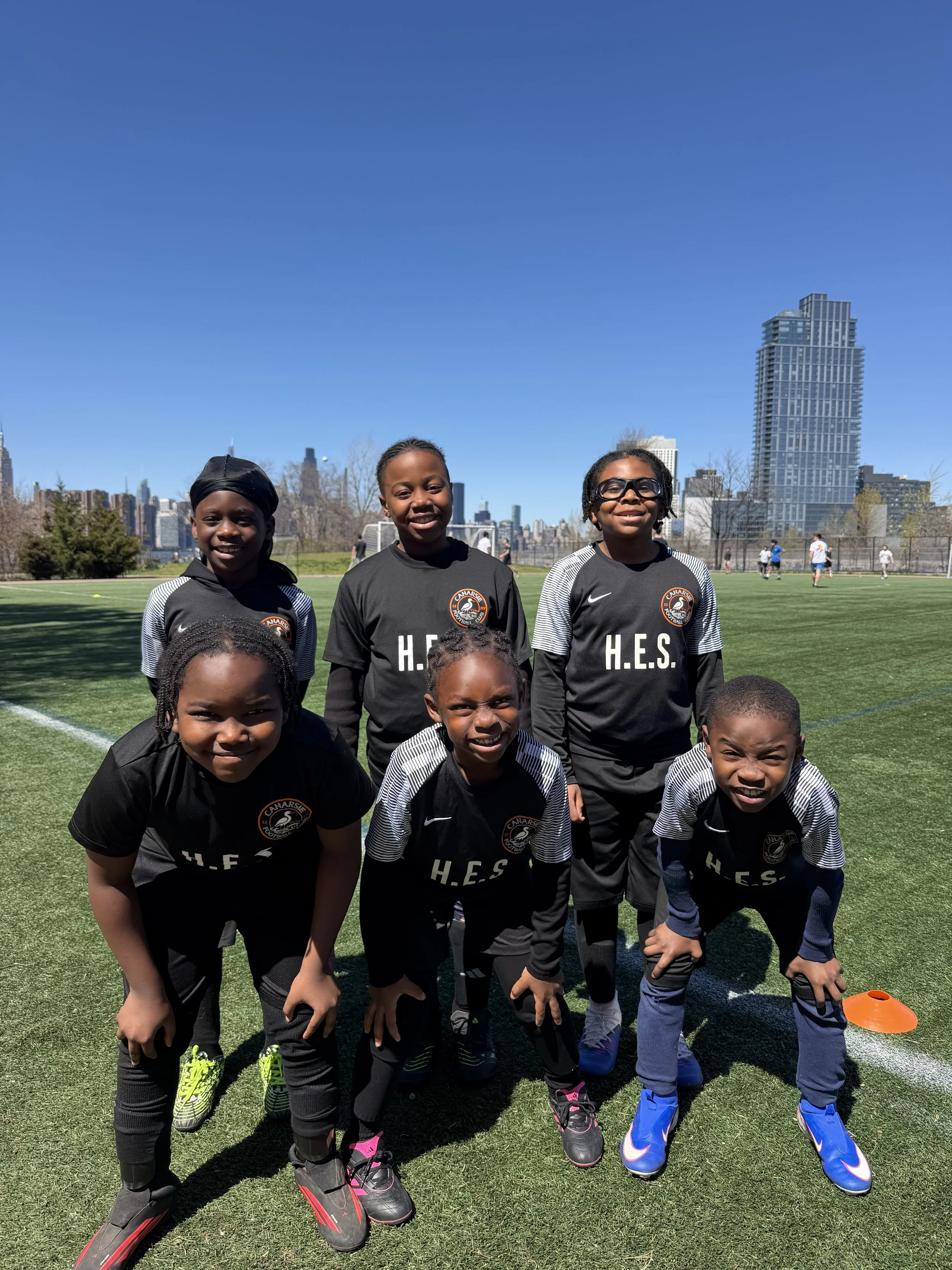 Group of young girls in black soccer uniforms with H.E.S. logo on a soccer field, with a city skyline featuring tall buildings in the background on a sunny day.