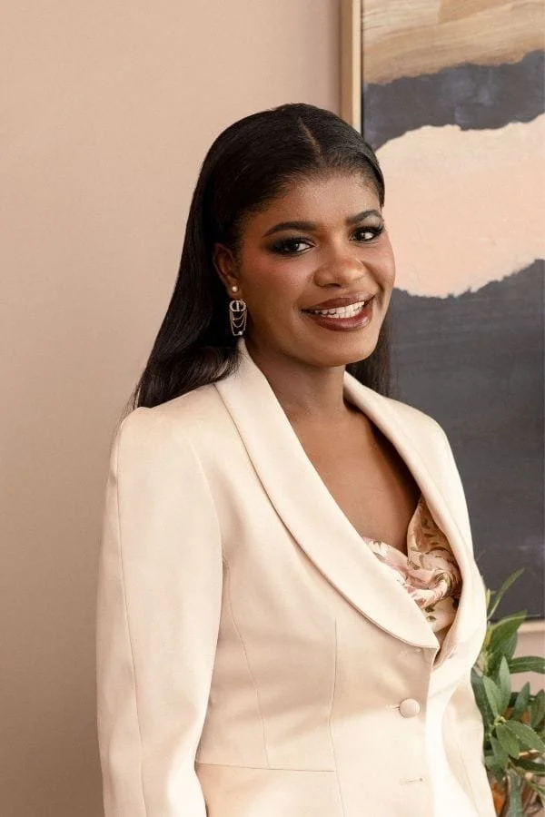 Portrait of Mercy de-Graft Savage, wearing a cream-colored blazer and earrings, standing indoors in front of a wall with a framed abstract painting and a plant.
