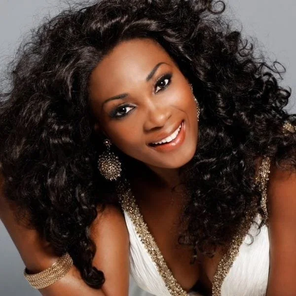 A woman with dark curly hair smiling at the camera, wearing large earrings and a white dress with gold embellishments.