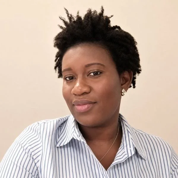 A woman with short natural curly hair, wearing a striped button-up shirt and earrings, smiling slightly.