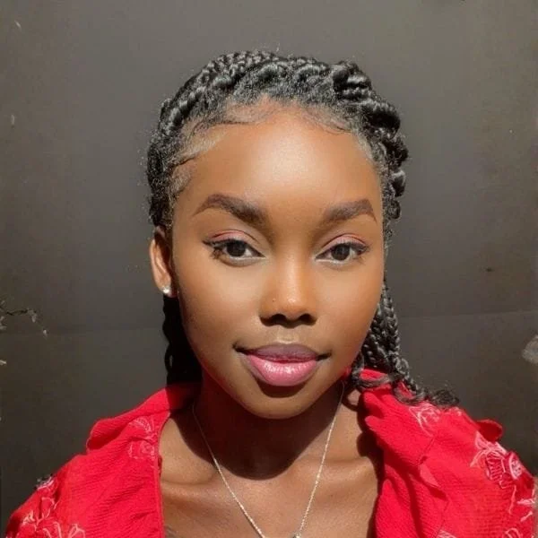 A young woman with braided hair, wearing a red top and silver jewelry, posing against a dark background.