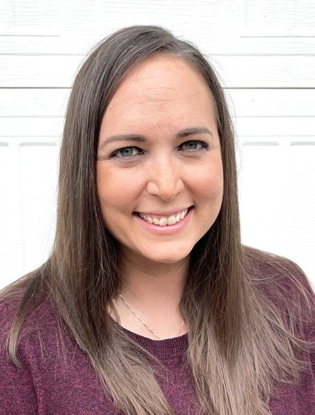 A smiling woman with long brown hair, wearing a maroon top, standing in front of a white wooden background.