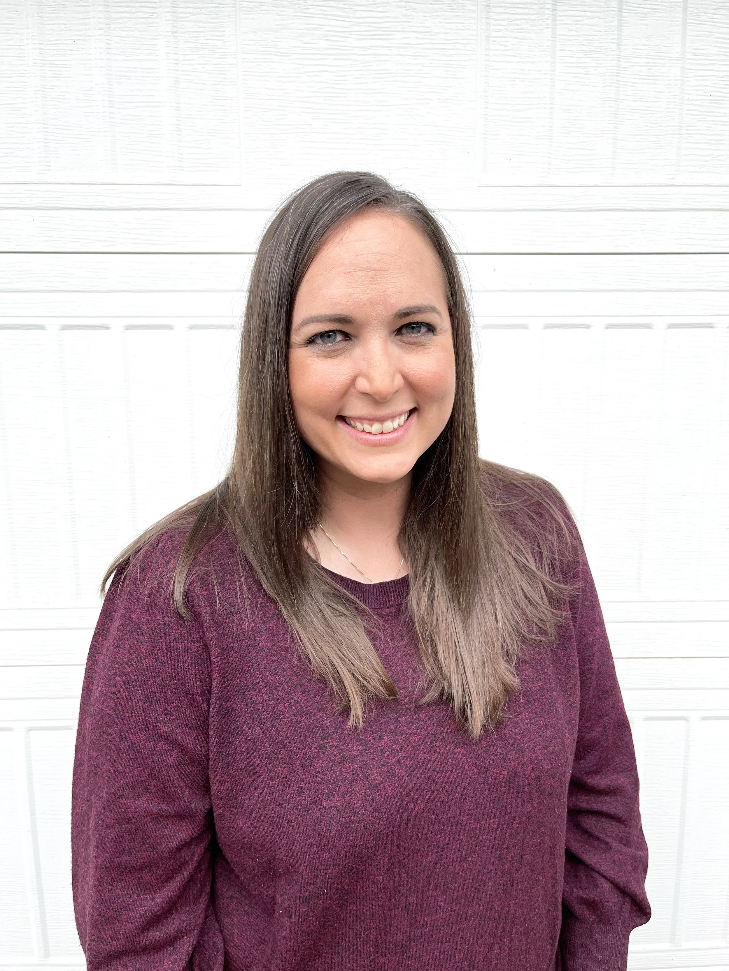A woman with long brown hair, blue eyes, and a bright smile, standing in front of a white wooden background, wearing a maroon sweater.