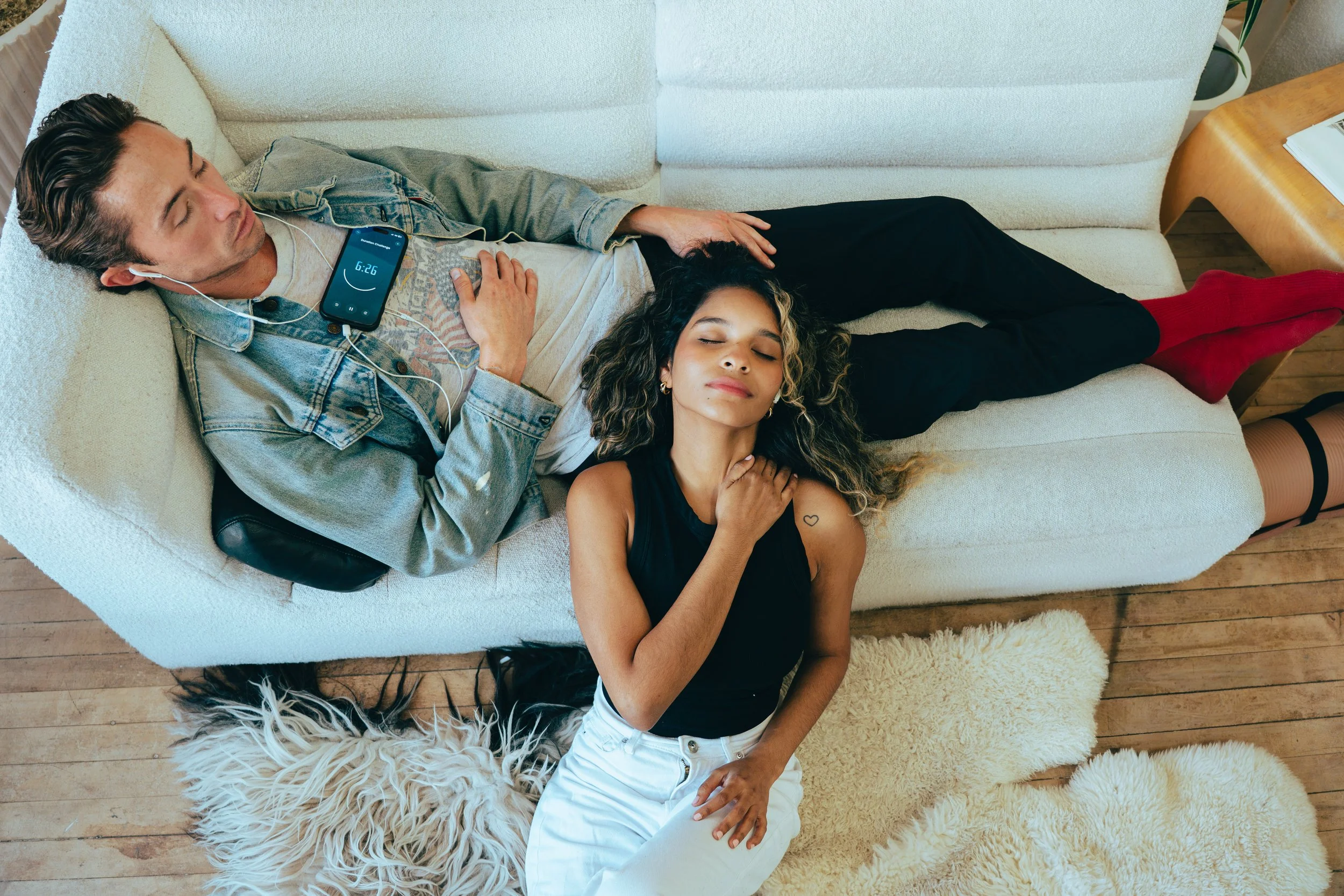 Man laying on white couch with headphones in and phone on his chest and a woman sitting on the floor next to the couch with her head leaning back on the man's lap. Both have closed eyes