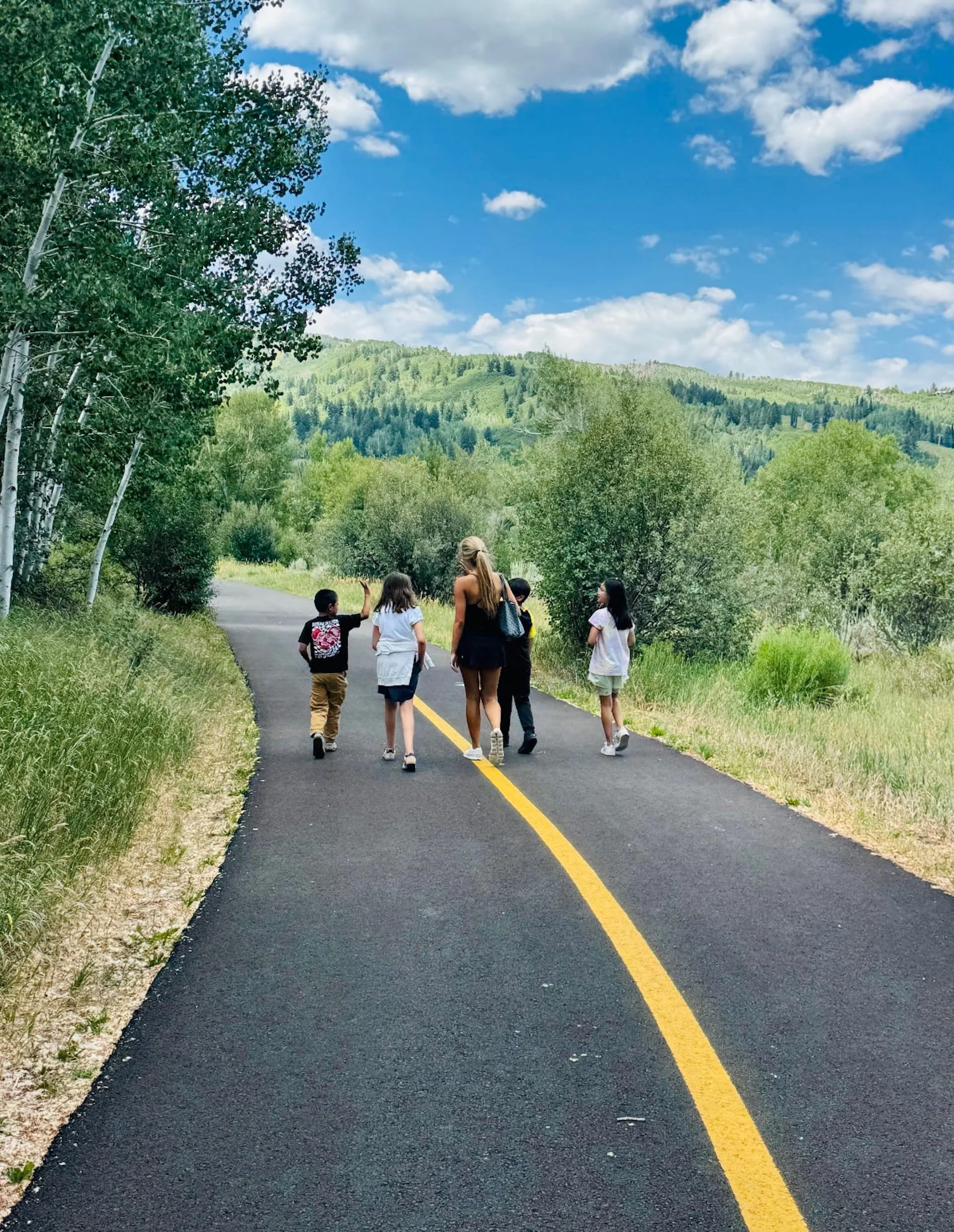A group of children and a woman walking on a paved road in a scenic outdoor area with green trees and hills, under a partly cloudy sky.