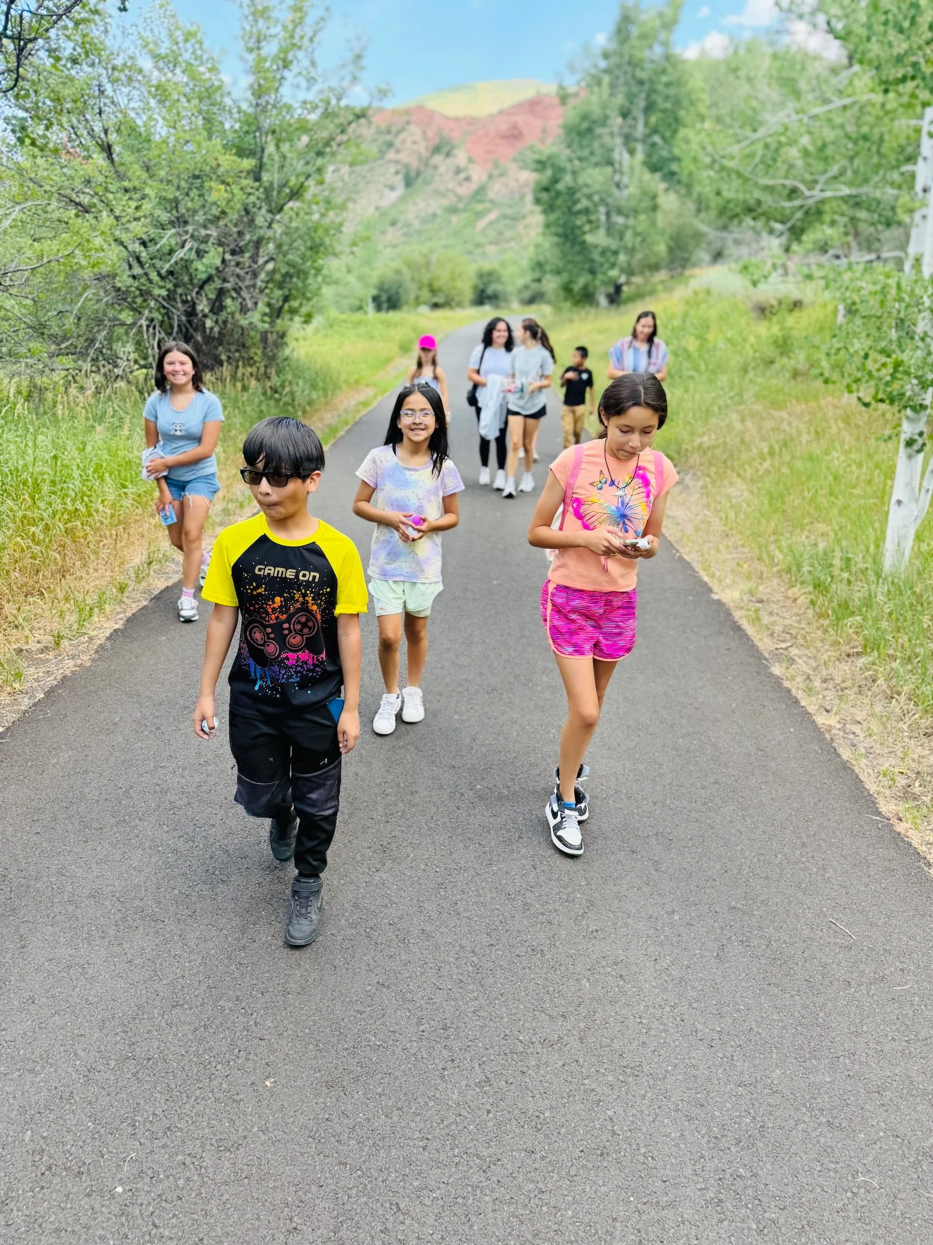 A group of children walking on a paved trail in a green, mountainous area.