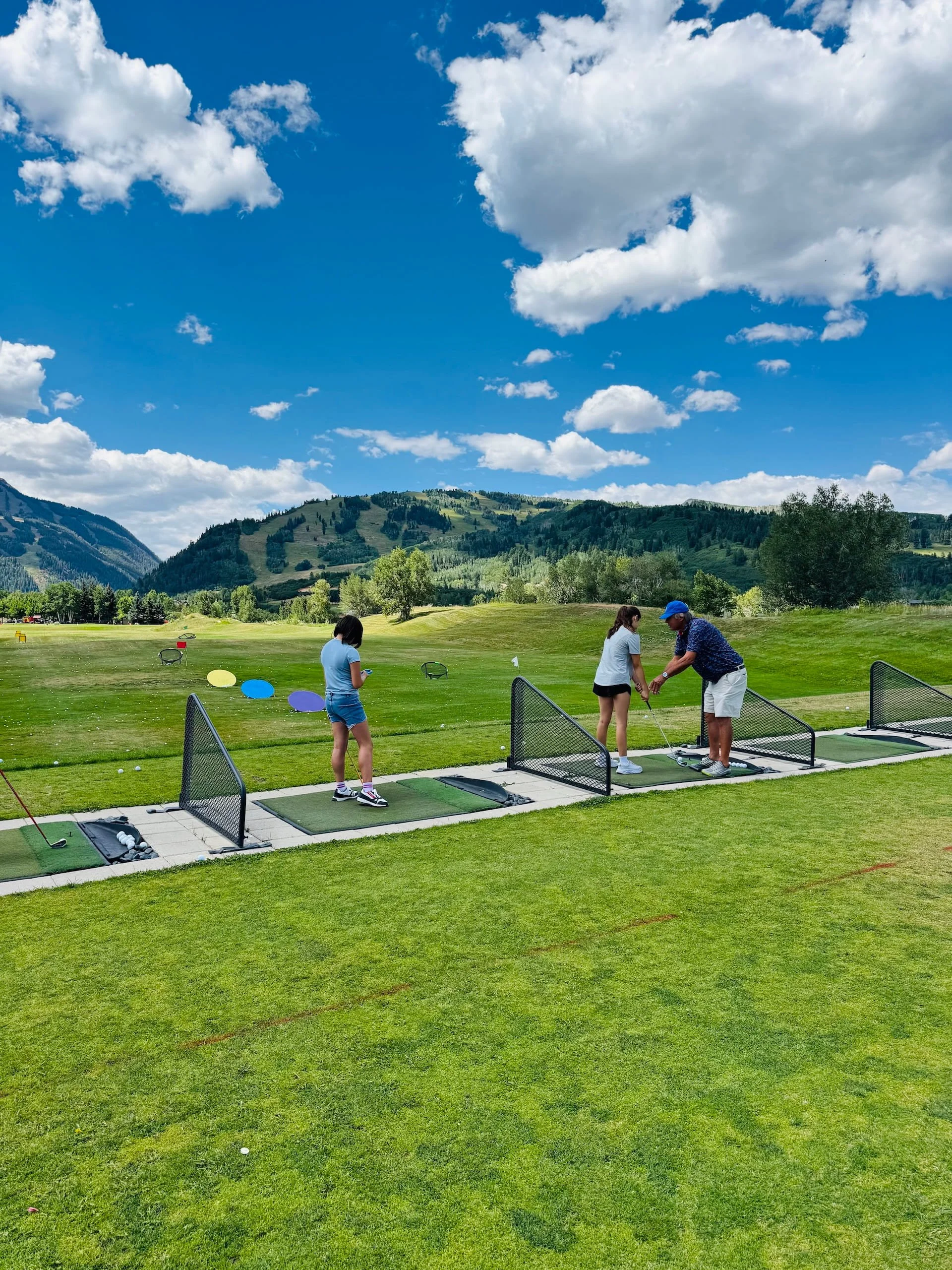 A group of people at a golf driving range practicing their swings under a bright blue sky with scattered clouds, with mountains and green trees in the background.