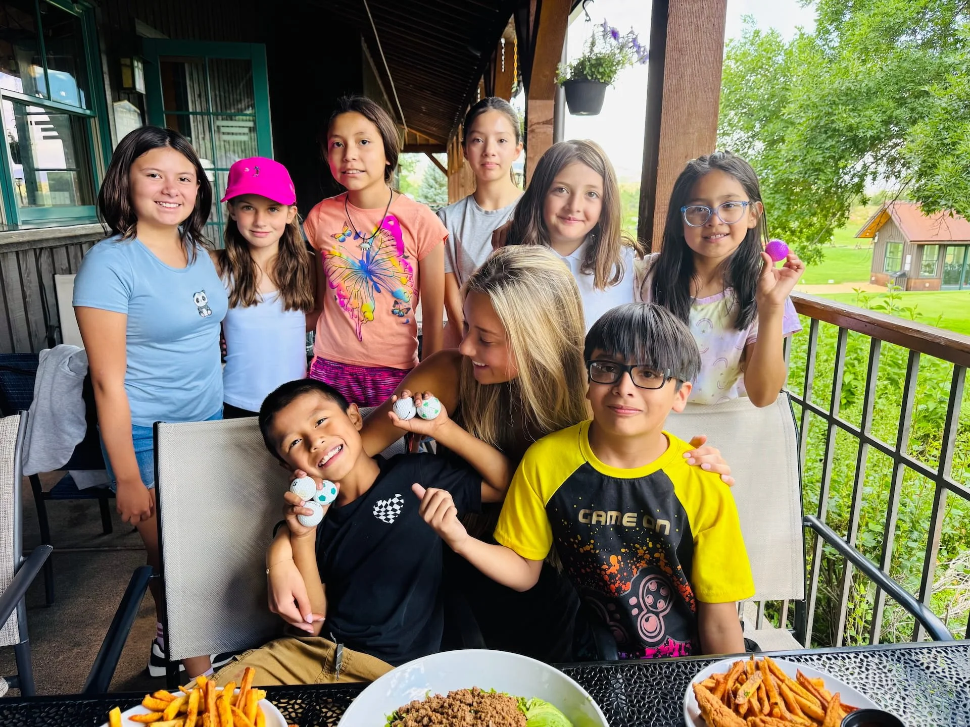 A group of kids, six girls and three boys, on a porch enjoying each other's company. Some are smiling and holding decorated eggs, with plates of food like fries and ground beef in the foreground. The background features lush green trees and small houses.