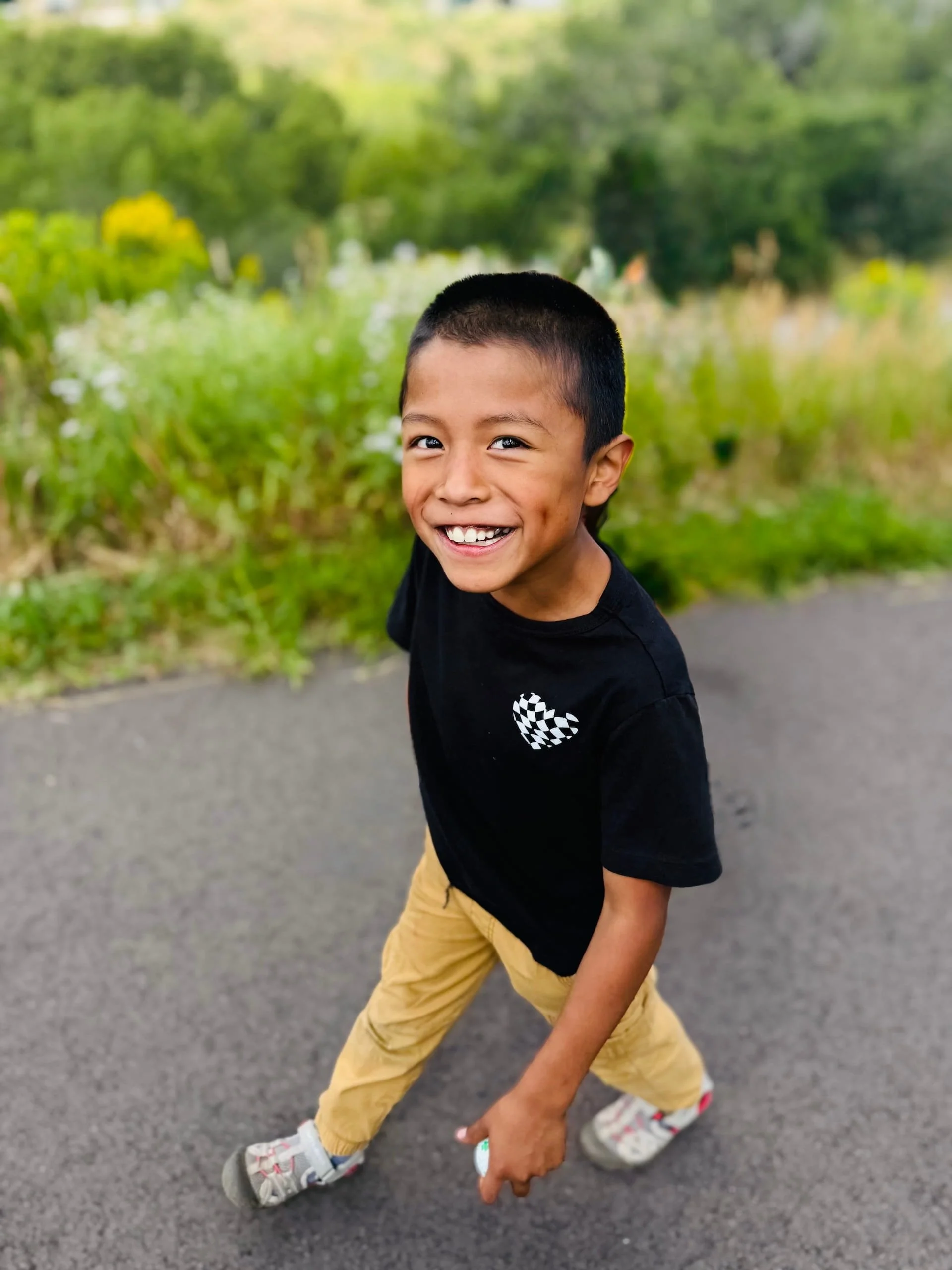 A young boy with short black hair smiling and looking at the camera, walking outdoors on a paved path with green trees and bushes in the background.