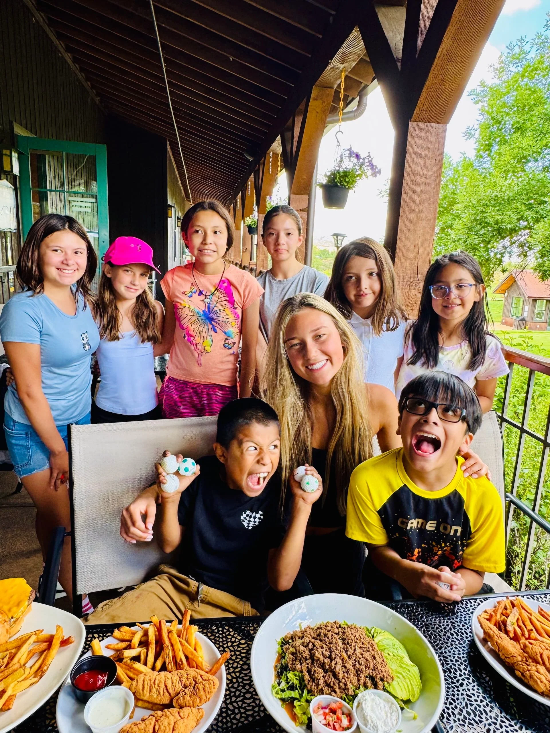 A group of children and a young woman are gathered on a covered porch with food, including fried chicken, fries, and salads, with green trees and a house in the background.