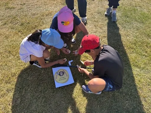 Three children crouch on a grassy field looking at a tablet, with a picture of a man and a yellow banner on the screen.