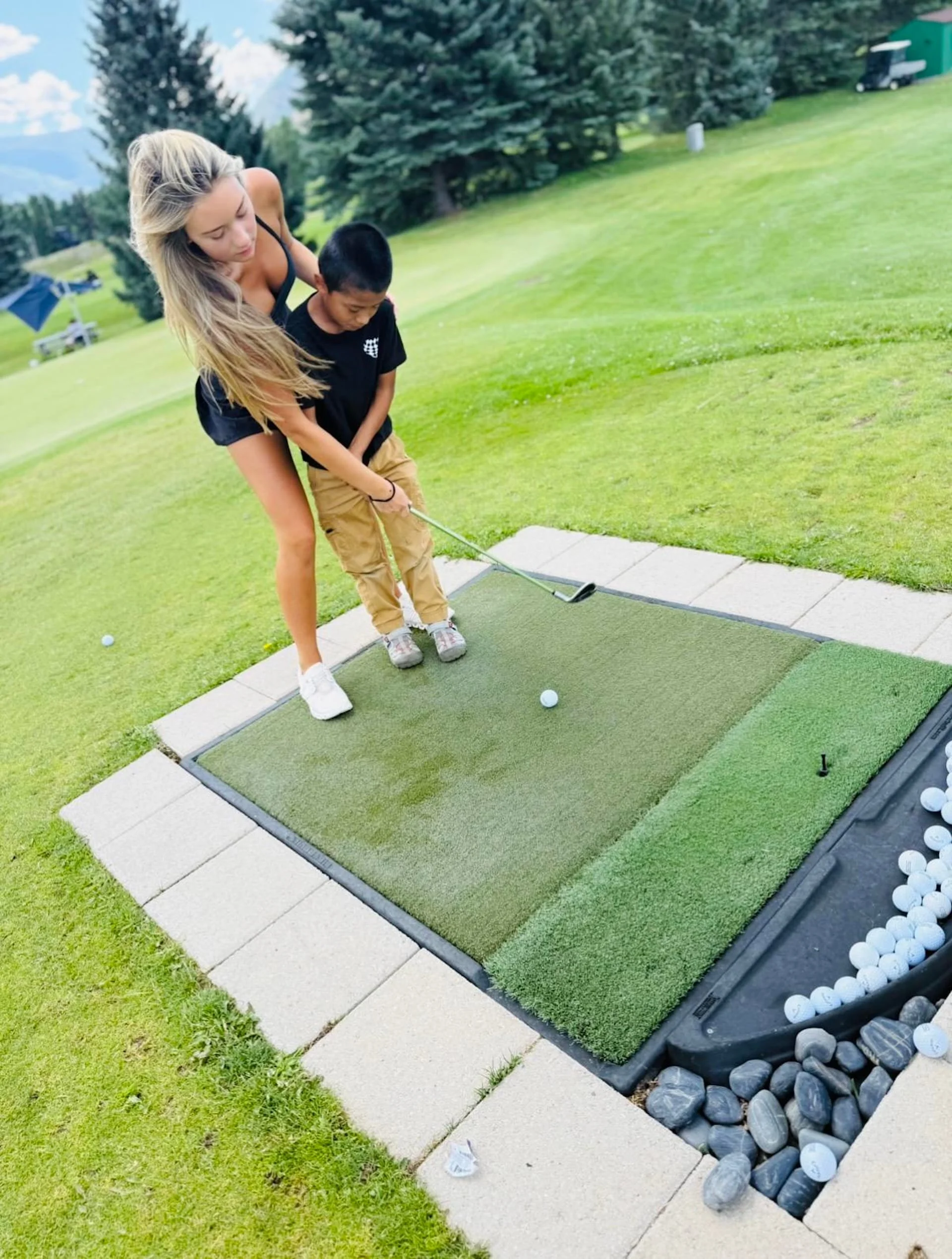 A woman and a young boy practicing golf on a practice mat outdoors surrounded by grass and trees. The woman is helping the boy with his golf swing.