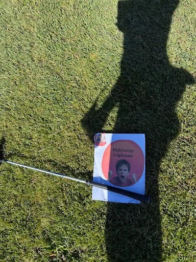 A golf club placed on the ground next to a sign that reads 'High Enemy Operations!' with a surprised child's face on it, cast in shadow on a grassy area.