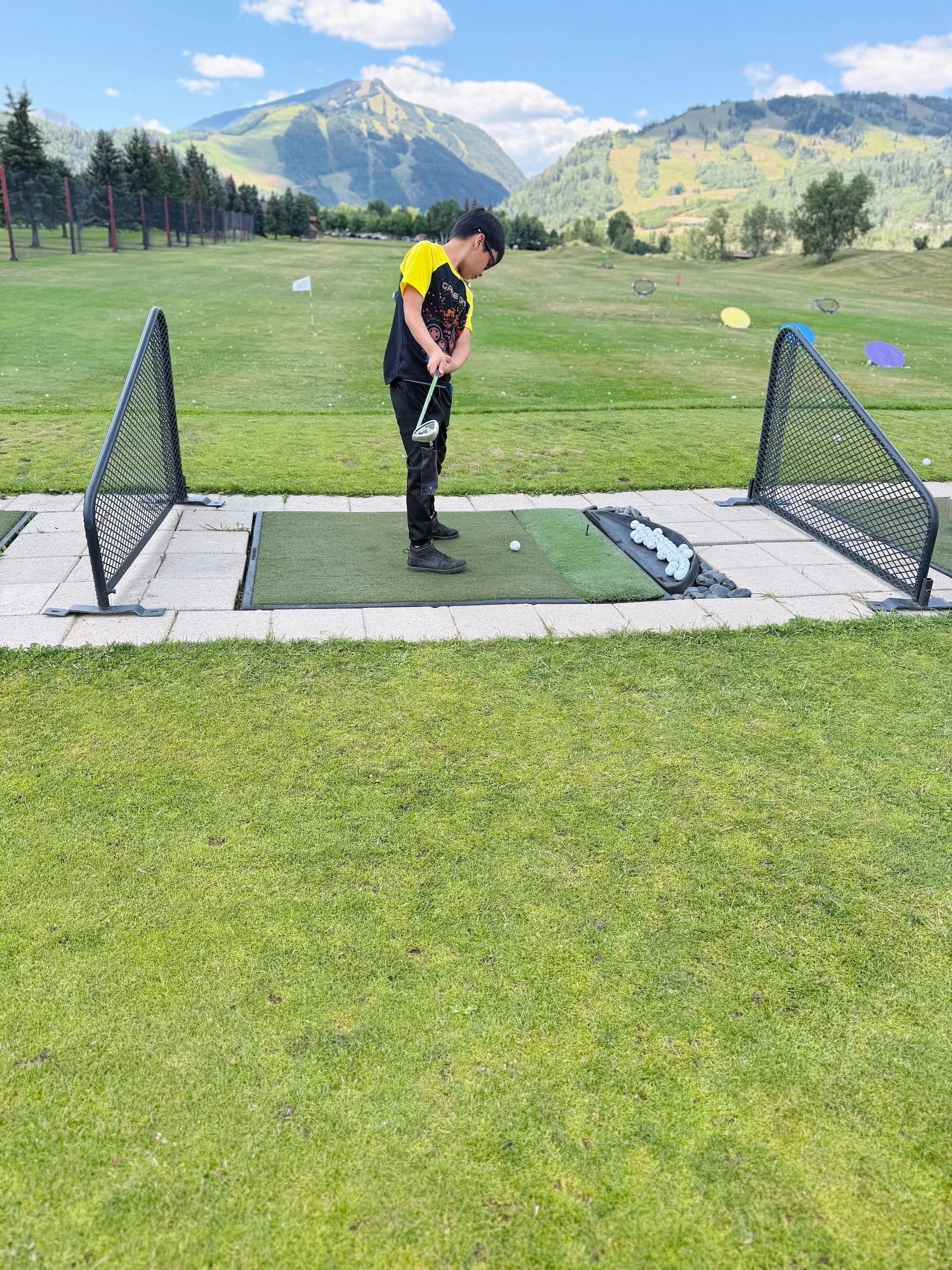 A young boy at a driving range practicing golf, surrounded by scenic mountains and a clear blue sky.