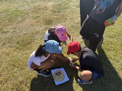 Three children and one adult sitting on grass, examining a medal and certificate. The children wear colorful hats, and one child has a bottle. The adult stands nearby holding a bottle.
