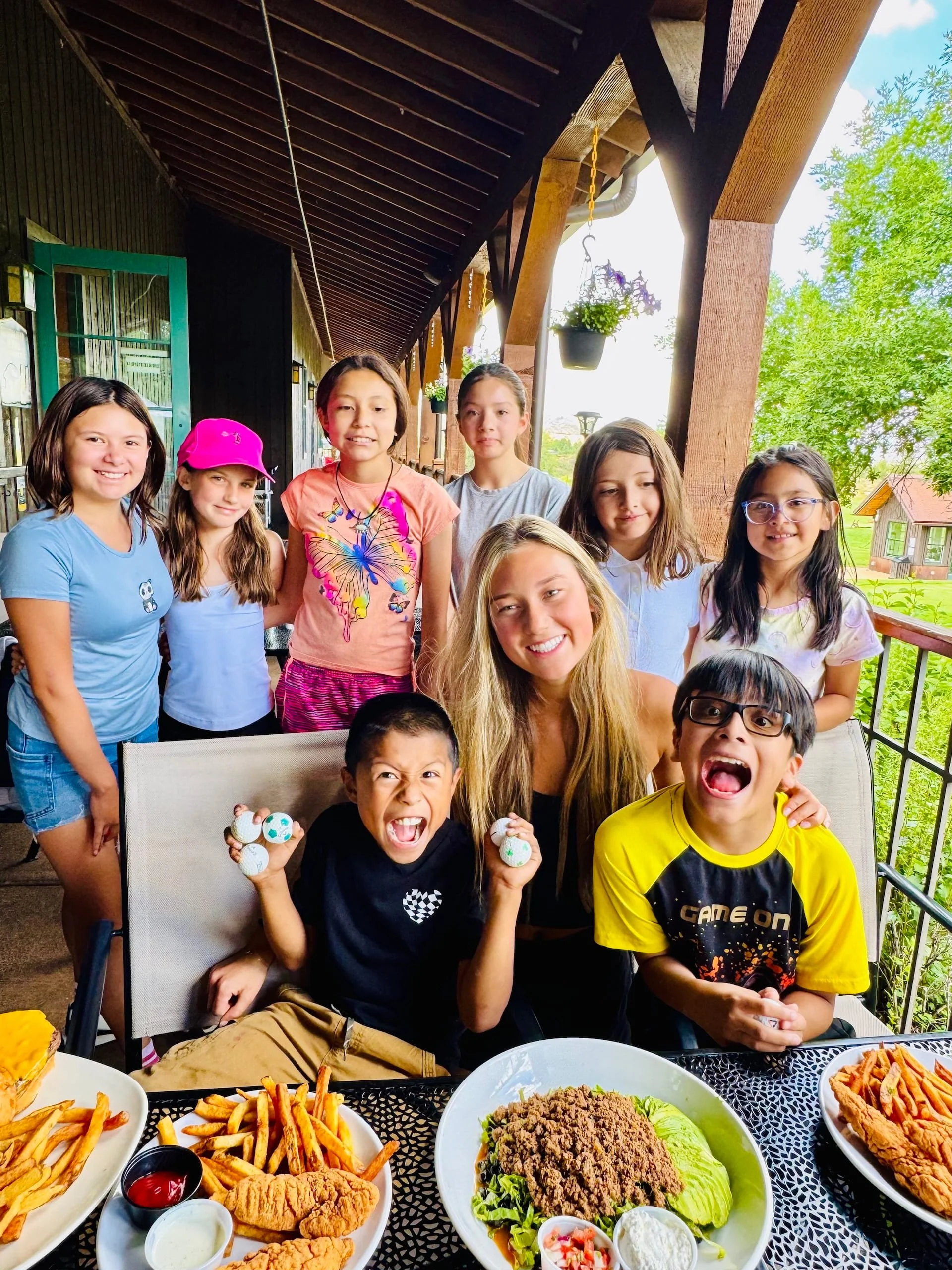 Group of children and a woman at a table with various dishes of food, including fries, fried chicken, and a large salad, on an outdoor porch with green trees and flowers in the background.