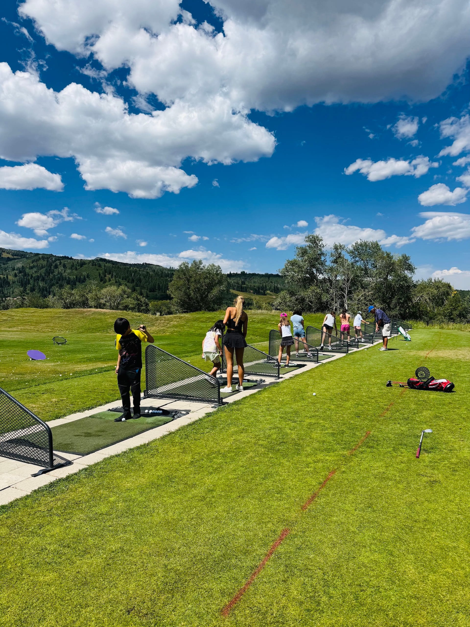 Children and adults practicing golf at an outdoor driving range with a scenic mountain landscape and blue sky with clouds in the background.