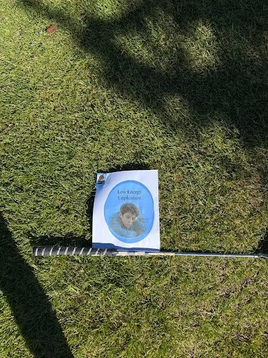 A book titled "Low Energy Supplements" lying on green grass beside a golf club, with shadows cast on the grass.