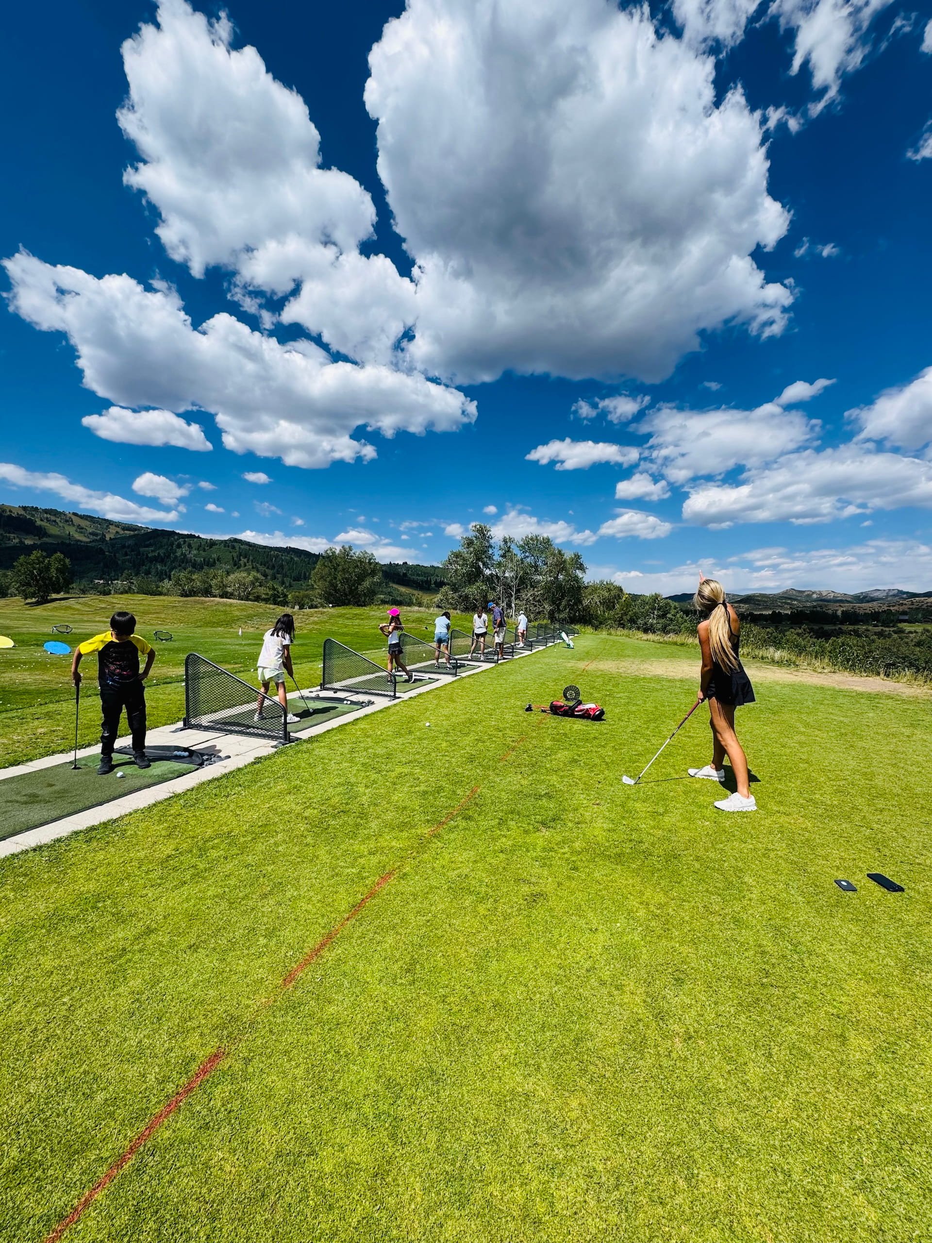 Children and a woman golfing on a sunny day at a golf practice area in a scenic outdoor setting with green grass, trees, and mountains in the background.