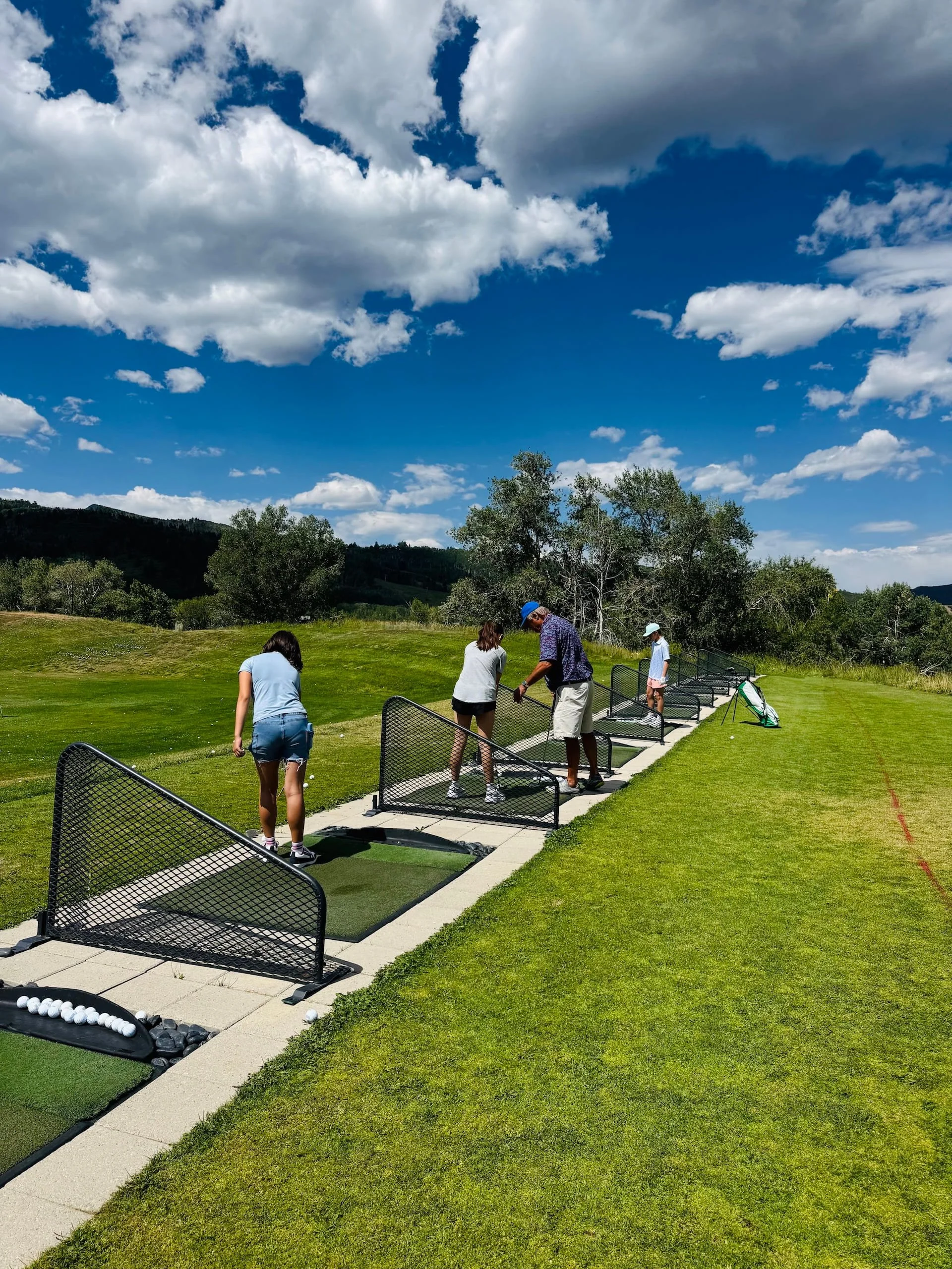A group of people at a golf driving range practicing their swings under a partly cloudy sky with green trees and hills in the background.