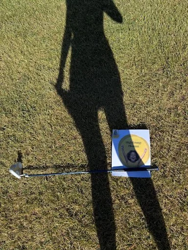 Shadow of a person on grass with a sign on the ground and a golf club lying next to it.