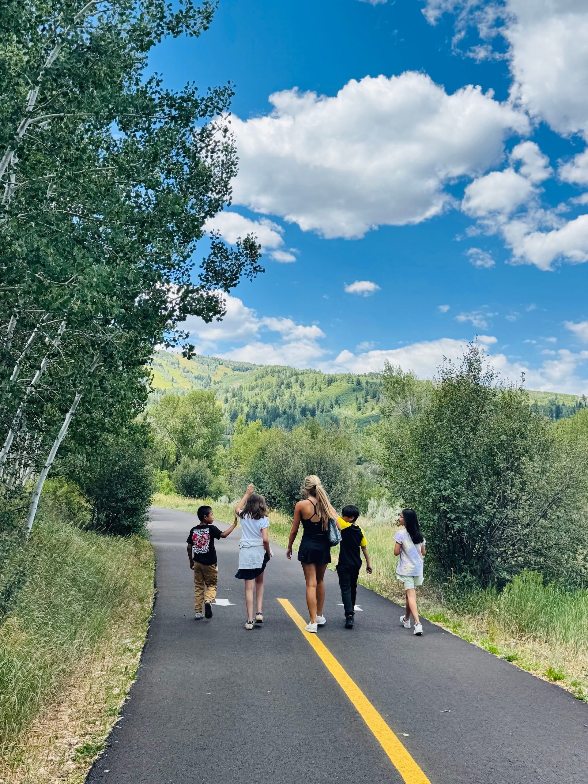 Group of children and a woman walking on a paved country road surrounded by greenery and trees, with hills and a partly cloudy sky in the background.