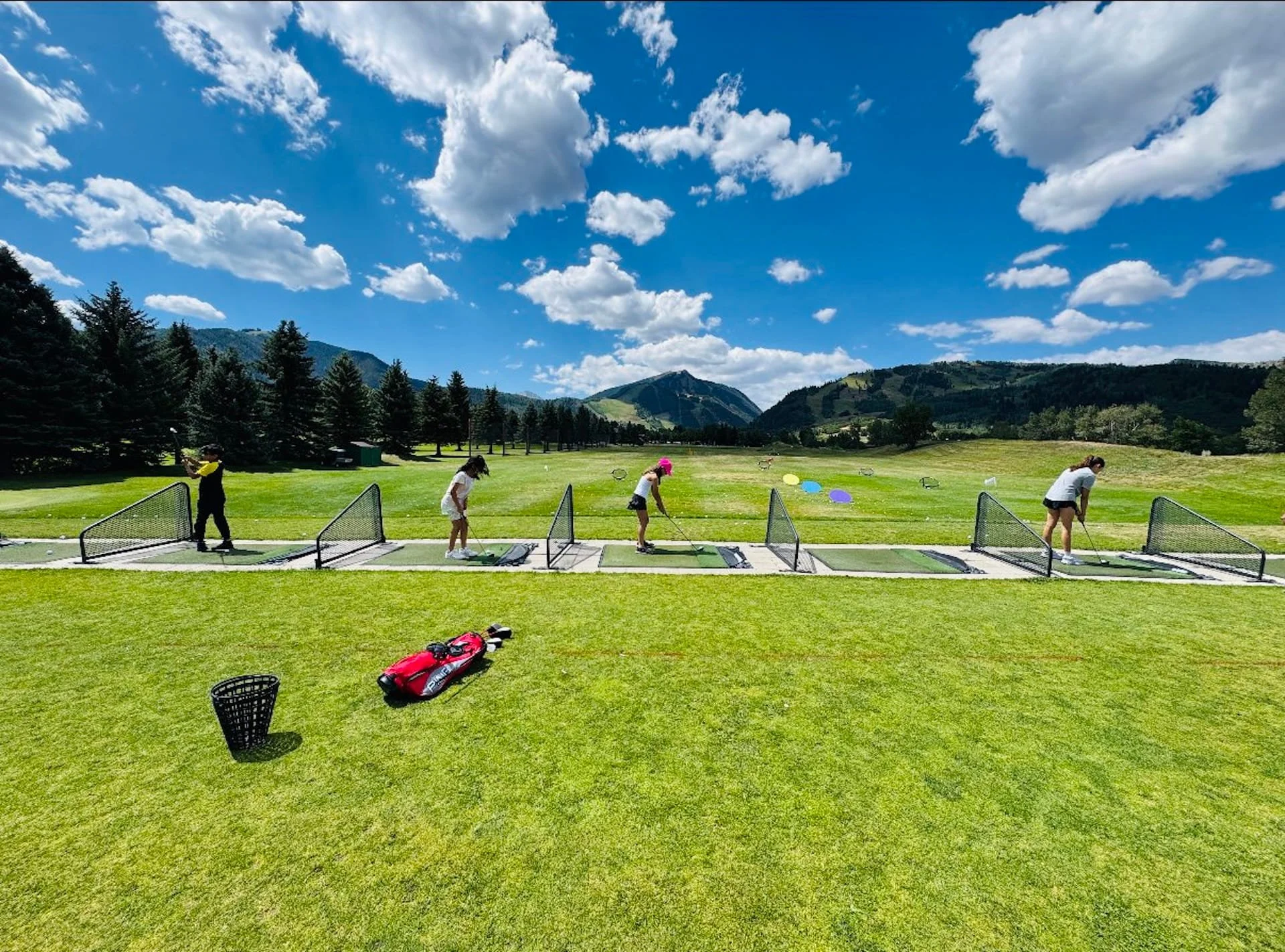 Four people practicing golf at an outdoor driving range with a mountainous landscape and blue sky with clouds in the background.
