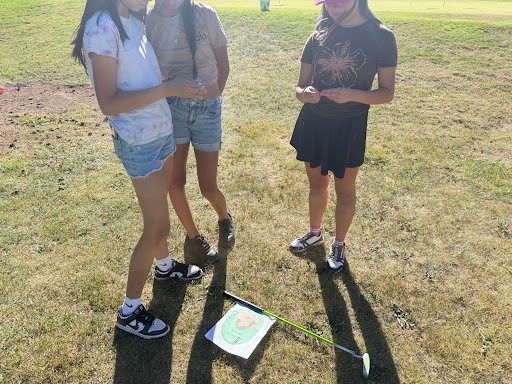 Three girls standing on grass in a park, showing a drawing on paper and a golf club.