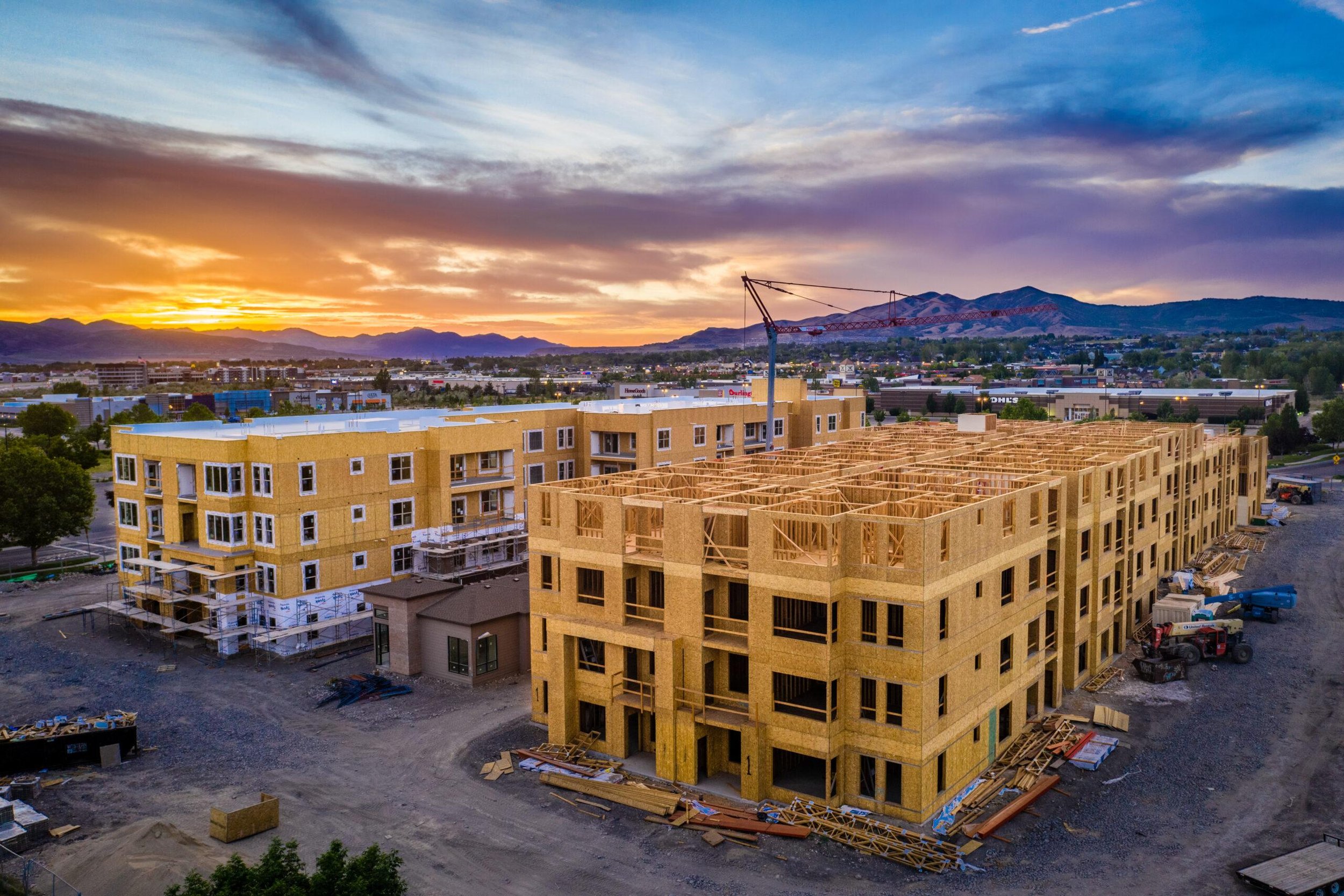 Construction site with two partially built wooden apartment buildings, a crane, and a cityscape with mountains in the background during sunset.