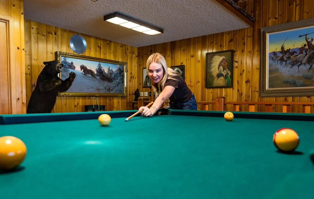 A woman playing pool in a room with wood-paneled walls decorated with wildlife paintings. A bear is standing on its hind legs with paws raised, positioned behind the woman, appearing as if it is reacting to her game.