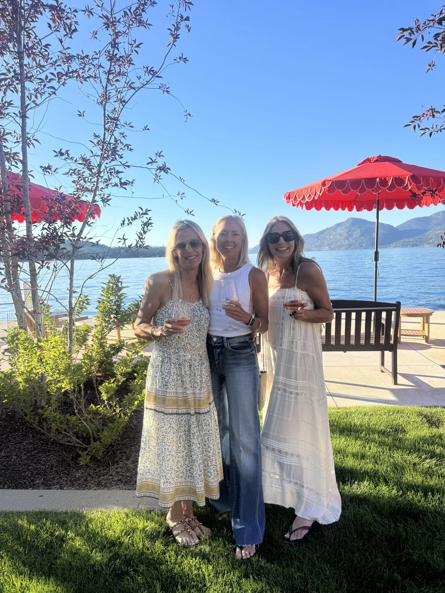 Three friends with drinks on the lakefront lawn under red umbrellas at The Retreat on Lake Pend Oreille