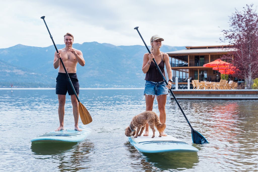 Two guests paddleboarding with a dog below the lodge at The Retreat on Lake Pend Oreille