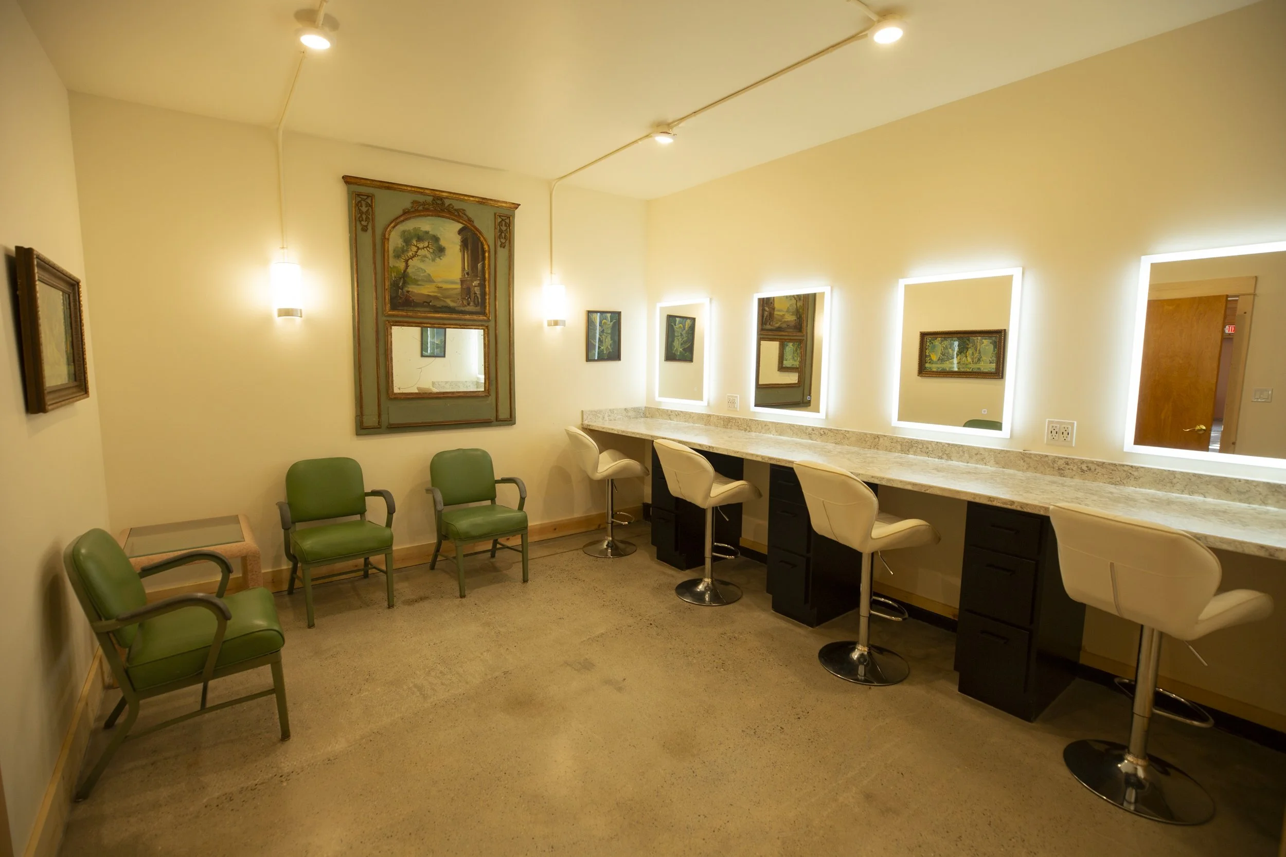 Interior of a small waiting or lounge area with green and beige chairs, a long granite counter with barstools, framed artwork on the walls, and recessed lighting.