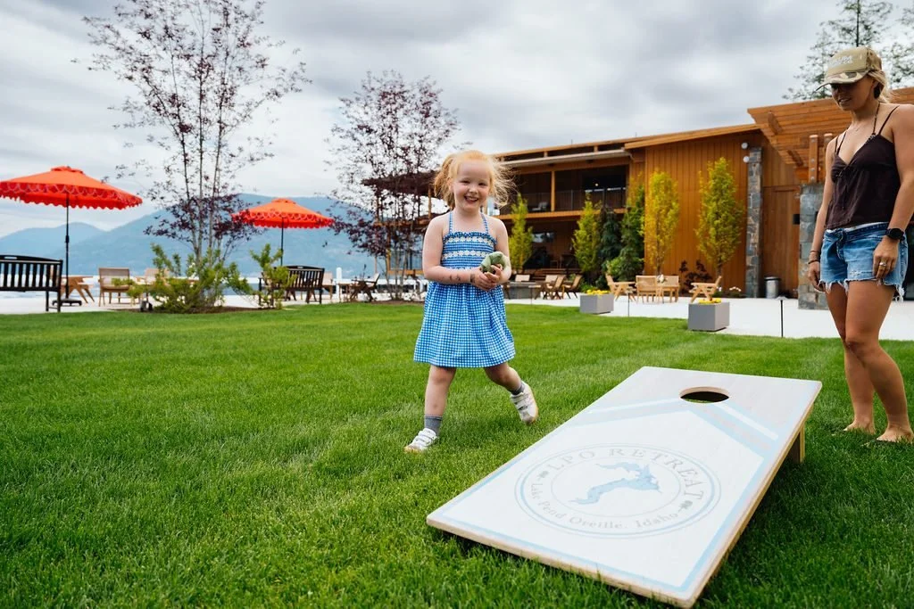 A girl in a blue gingham dress playing cornhole with an adult woman in a black tank top and denim shorts on a grassy lawn. There are orange umbrellas, trees, and a building with outdoor seating in the background.