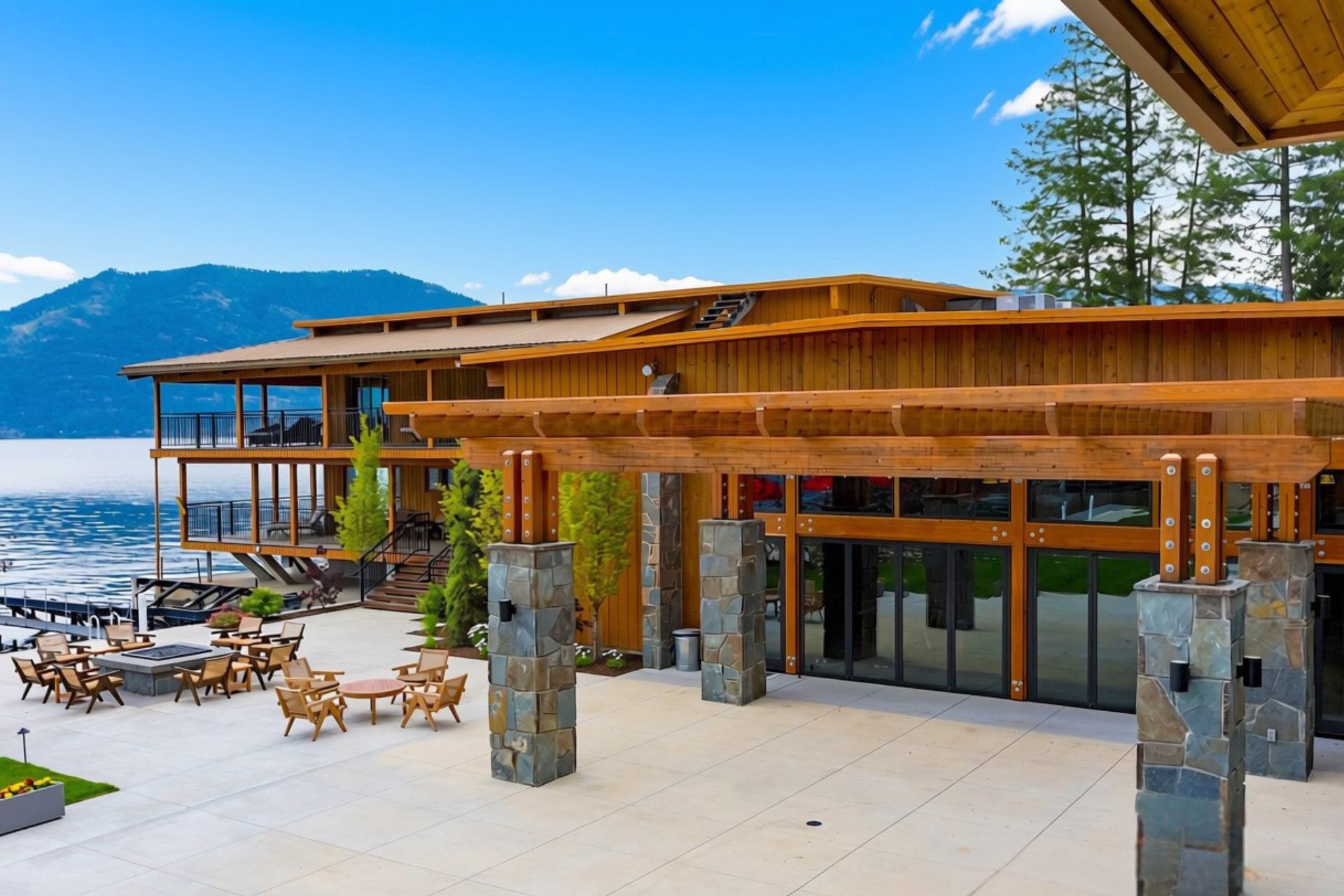Modern multi-story lakeside building made of wood and stone with outdoor patio and furniture, overlooking a mountain and lake under a clear blue sky.